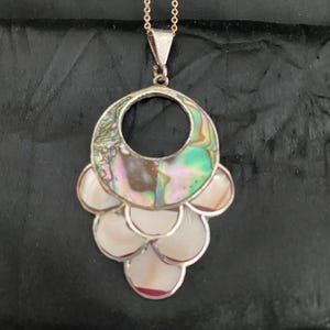 Pu&ograve; includere: Una collana d'argento con un grande ciondolo in madreperla di abalone iridescente. Il ciondolo ha la forma di un cerchio con un cerchio pi&ugrave; piccolo tagliato al centro. Il ciondolo &egrave; circondato da pezzi pi&ugrave; piccoli di madreperla di abalone.