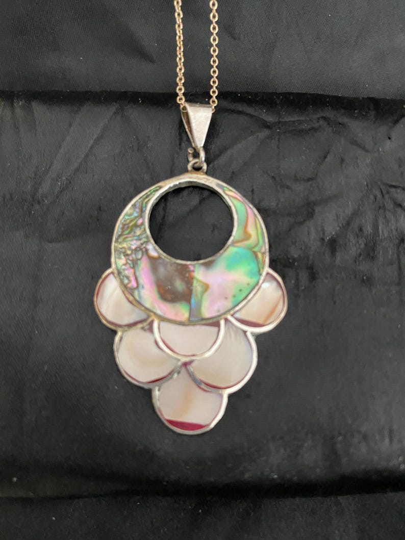 Pu&ograve; includere: Una collana d'argento con un grande ciondolo in madreperla di abalone iridescente. Il ciondolo ha la forma di un cerchio con un cerchio pi&ugrave; piccolo tagliato al centro. Il ciondolo &egrave; circondato da pezzi pi&ugrave; piccoli di madreperla di abalone.