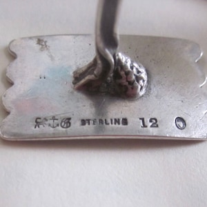 May include: Close-up of the bottom of a silver figurine. The base is marked with "Sterling 12" and a small anchor symbol.