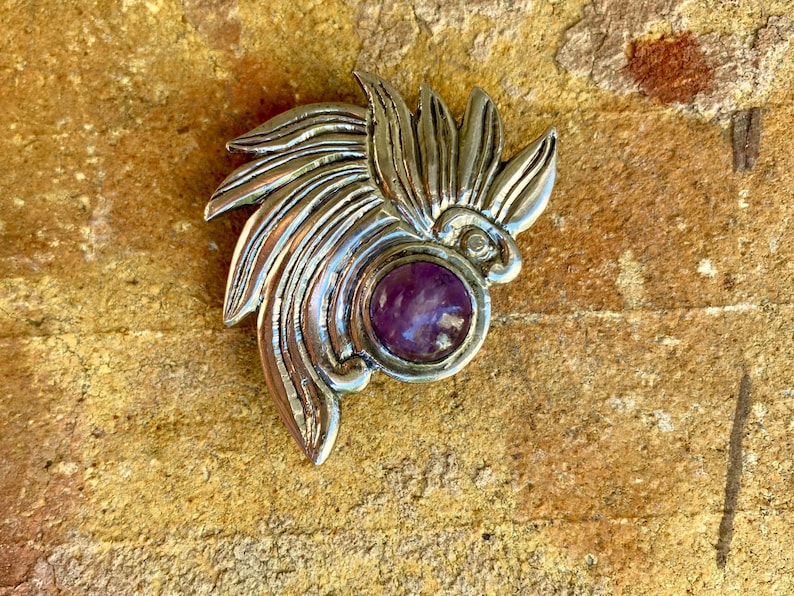 May include: A silver-toned metal brooch with a stylized feather design and a central, round, purple gemstone. The brooch is set against a textured, tan-colored surface, showcasing its intricate details and craftsmanship.