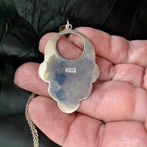 Pu&ograve; includere: Una collana con un ciondolo in argento, grande, ornato e traforato. Il ciondolo ha la forma di un fiore stilizzato con un'apertura circolare al centro. Il retro del ciondolo &egrave; timbrato con la parola "Messico".