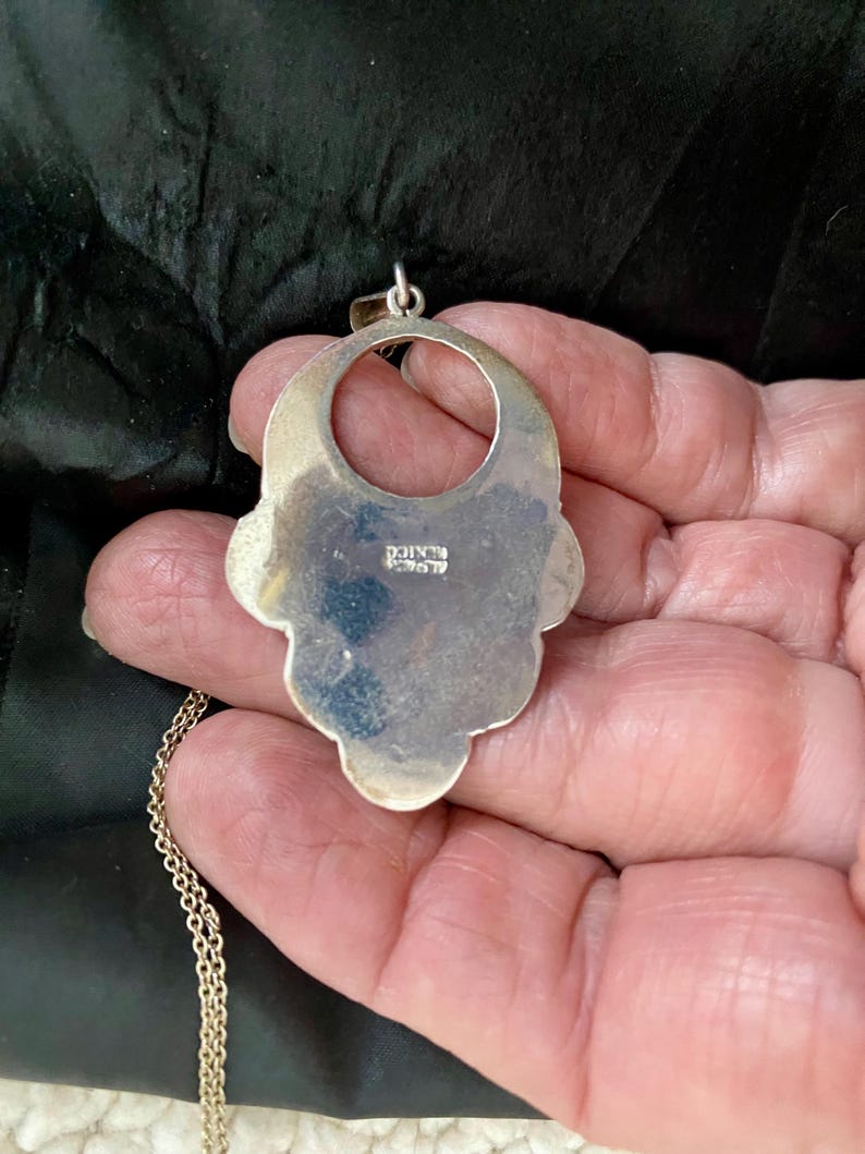 Pu&ograve; includere: Una collana con un ciondolo in argento, grande, ornato e traforato. Il ciondolo ha la forma di un fiore stilizzato con un'apertura circolare al centro. Il retro del ciondolo &egrave; timbrato con la parola "Messico".
