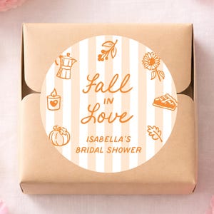 May include: A square brown cardboard box featuring a circular sticker with a white and light orange vertical striped background. The sticker displays the orange text Fall In Love and Isabella's Bridal Shower, surrounded by icons of a coffee pot, sunflower, candle, pumpkin, pie, and leaf.
