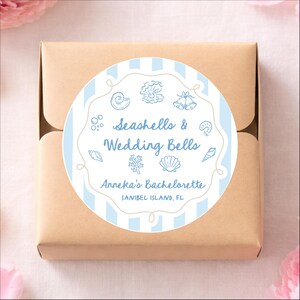 May include: A wrapped gift box with a round sticker featuring a blue and white striped background. The sticker has seashell and wedding bell illustrations, with the text "Seashells & Wedding Bells" and "Anneka's Bachelorette, Sanibel Island, FL."