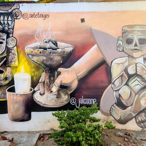May include: A mural featuring ancient figures and a hand holding a smoking vessel. The artwork includes a candle, a small statue, and the text "@jakcoone" and "@artetinyjei". The color palette is earthy, with browns, tans, and hints of red.