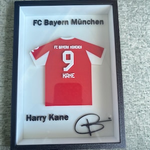 Pode incluir: Obra de arte emoldurada com uma camisa vermelha e branca do FC Bayern Munique com o número 9 e o nome KANE. A moldura inclui o texto "FC Bayern München" e "Harry Kane" com uma assinatura.