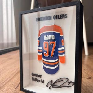 Puede incluir: Obra de arte enmarcada con una camiseta de hockey de los Edmonton Oilers. La camiseta es azul, naranja y blanca, con el nombre "McDavid" y el número "97". El marco incluye el texto "Edmonton Oilers" y una firma.
