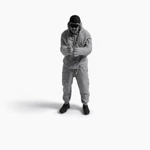 Puede incluir: Una figura en miniatura en escala de grises de una persona con sudadera con capucha, pantalones de chándal, gafas de sol y gorra. La figura está de pie con las manos levantadas. La figura lleva zapatos negros.