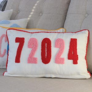 Könnte beinhalten: Eine Sammlung weißer Zierkissen mit bunten Stickereien. Ein Kissen zeigt die Zahl "72024" in Rot und Rosa. Andere Kissen zeigen einen Regenbogen, Wolken und ein Auto.