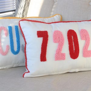 Op de afbeelding: Twee witte rechthoekige kussens met kleurrijk geborduurde tekst. Eén kussen heeft "CU" in roze en blauw. De andere heeft "72024" in rood en roze. Beide hebben een gekleurde rand.
