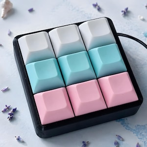 Pode incluir: Um teclado macro mecânico preto com nove teclas quadradas dispostas numa grelha de três por três. A fila superior é branca, a fila central é azul-clara e a fila inferior é cor-de-rosa clara. O texto Ocean kiss está impresso acima do teclado.