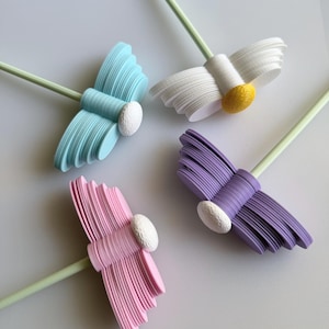 Könnte beinhalten: Vier dekorative Gänseblümchen-Dekorationen in Pastellfarben. Die Blütenblätter sind hellblau, weiß, rosa und lila. Jede Blume hat eine weiße oder gelbe Mitte und einen hellgrünen Stiel. Die Blumen sind auf einer weißen Oberfläche angeordnet.