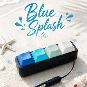 Könnte beinhalten: Ein schwarzes Makro-Pad mit vier Tasten für mechanische Tastaturen mit einem Farbverlauf der Tastenkappen von Dunkelblau bis Weiß. Über dem Gerät steht der Text Blue Splash, das mit einem schwarzen Kabel mit zylindrischem Stecker verbunden ist.