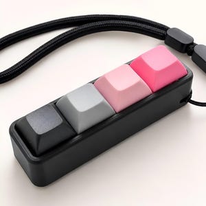 May include: A black rectangular mechanical keyboard accessory featuring four individual keycaps in a gradient colour scheme of black, grey, light pink, and dark pink. The device is attached to a black woven lanyard with a plastic adjustment bead.