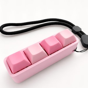 Könnte beinhalten: Ein Satz von vier rosa Tastaturkappen in einem rosa Tablett, mit einer schwarzen Handschlaufe. Die Tastenkappen sind in verschiedenen Rosatönen gehalten. Der Hintergrund ist weiß.