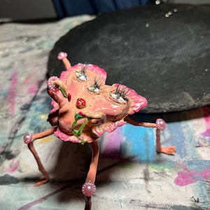 May include: A whimsical, handmade sculpture featuring a pink, heart-shaped face with multiple eyes, long eyelashes, and a red mouth. The sculpture has thin, pink legs with pearl accents and is posed on a colorful surface.