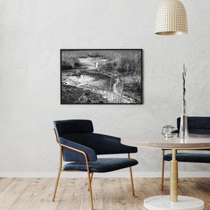 Może przedstawiać: Czarno-biała oprawiona fotografia przedstawiająca scenę z bagna. Fotografia przedstawia wijący się strumień przez wysokie trzciny i trawy. Zdjęcie wisi nad stołem i krzesłami.