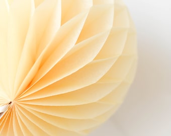 May include: A close-up of a large, round, cream-colored paper honeycomb ball. The ball is made of many layers of paper that are folded and overlapped to create a honeycomb pattern.