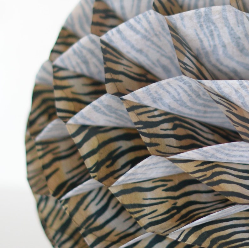 May include: A close-up of a decorative paper ball with a tiger stripe pattern. The ball is made of multiple layers of paper, each with a different color and pattern. The ball is a light brown color with black stripes.