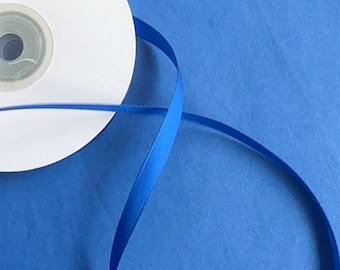 May include: A spool of vibrant royal blue satin ribbon with a smooth, reflective finish. The ribbon is wound around a white circular plastic core, with a single length of the blue ribbon extending outward and curving into a loose, open loop.