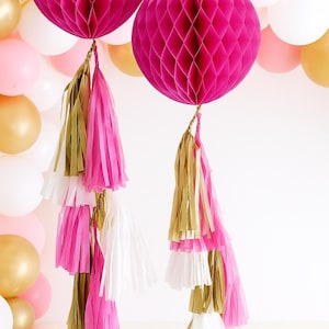 May include: Two fuchsia honeycomb paper decorations with gold and white tassels hang in front of a backdrop of gold, pink, and white balloons. These festive decorations are perfect for parties and celebrations.