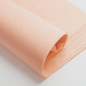 May include: A stack of thin, semi-transparent peach-colored tissue paper sheets. The top sheet is partially rolled over, revealing the delicate, lightweight texture and consistent light orange hue of the paper stack, which is neatly aligned.