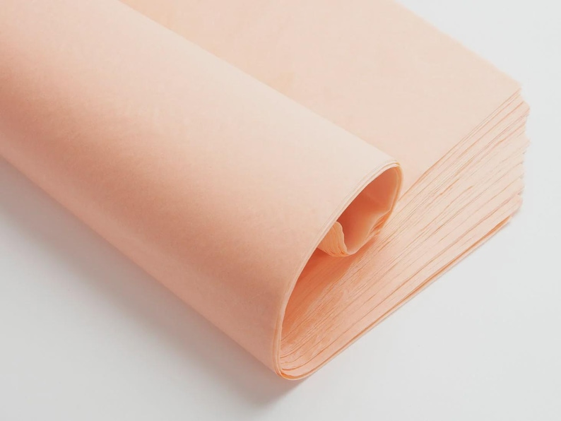 May include: A stack of thin, semi-transparent peach-colored tissue paper sheets. The top sheet is partially rolled over, revealing the delicate, lightweight texture and consistent light orange hue of the paper stack, which is neatly aligned.