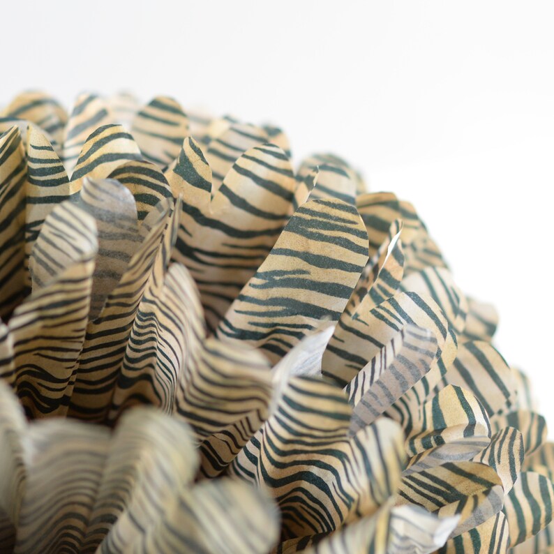 May include: Close-up of a brown and black striped tissue paper pom-pom. The paper is crinkled and folded to create a fluffy, round shape.