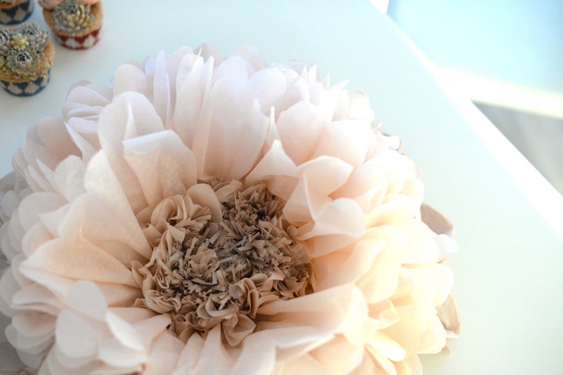 May include: A large, pale pink tissue paper flower with a brown center. The flower is made of many layers of tissue paper and has a delicate, feminine look.