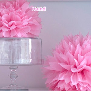 K&ouml;nnte beinhalten: Zwei rosa Seidenpapier-Pompons, einer rund und einer spitz, auf einem wei&szlig;en Regal. Der runde Pompon befindet sich auf einem gl&auml;sernen Kuchenteller.