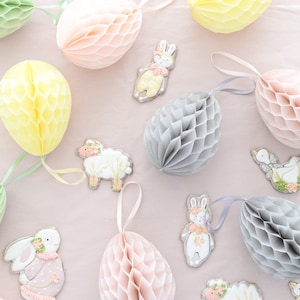 Op de afbeelding: Een verzameling pastelkleurige honingraat-weefselpapierdecoraties in roze, geel en grijs. De decoraties hangen aan een lichtroze achtergrond. Er zijn ook verschillende koekjesuitsteekvormen van konijnen en lammetjes.