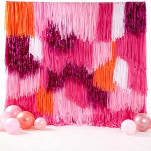 May include: A vibrant party backdrop featuring a cascade of pink, orange, and white fringe. The backdrop has a layered design with varying shades of pink and metallic accents. Several pink balloons are scattered in front of the backdrop.