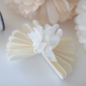 May include: A white paper flower with a layered, fan-like design. The flower is made from multiple layers of white paper that are folded and layered to create a three-dimensional effect. The flower is a popular craft project and can be used for a variety of purposes, such as decorating a home or adding a touch of elegance to a special event.