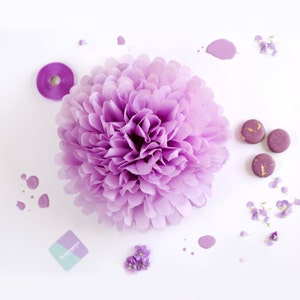 Op de afbeelding: Een grote, lichtpaarse papieren bloem met gelaagde bloemblaadjes staat centraal in de afbeelding. Paarse macarons met gouden accenten, verfspatten, een rol lint en kleine paarse bloemen zijn te zien op een witte achtergrond.