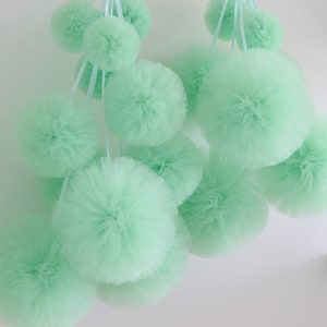 May include: A collection of light green tulle pom-poms of various sizes hanging from a white ceiling. The pom-poms are made of soft, fluffy tulle and are perfect for adding a touch of whimsy to any room.