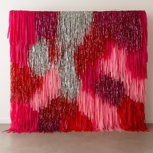 Pode incluir: Uma vibrante tapeçaria de parede composta por materiais em camadas e com franjas em tons de rosa, vermelho e prata. O design apresenta uma mistura de texturas sólidas e metálicas, criando um efeito visual dinâmico. A peça é provavelmente usada para fins decorativos.