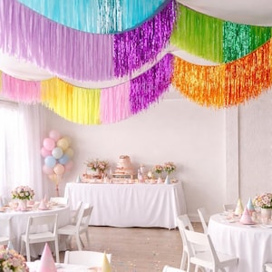 Könnte beinhalten: Festliche Partydekoration mit farbenfrohen Girlanden. Die Decke ist mit blau, lila, grün, orange und rosa Fransen geschmückt. Tische sind mit weißen Tischdecken, Partyhüten und Blumengestecken dekoriert, was eine festliche Atmosphäre schafft.