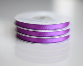 May include: A stack of three spools of vibrant purple satin ribbon. Each spool features a smooth, shiny texture and a consistent width, wound tightly around a white circular core. The ribbon displays a deep, saturated violet hue throughout.