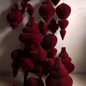 Op de afbeelding: Een verzameling dieprode papieren decoraties in honingraatvorm in verschillende vormen, waaronder bollen, druppels en geometrische vormen. Sommige hangen, andere zijn op een stoel geplaatst, wat een feestelijke en artistieke presentatie creëert. De oppervlakken zijn getextureerd.