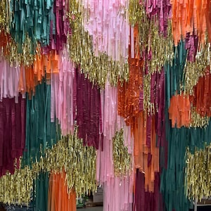 May include: A vibrant backdrop composed of hanging strips in shades of pink, orange, green, and burgundy, with gold tinsel accents. The strips are suspended from a metal rod, creating a textured, celebratory look. Suitable for parties and events.