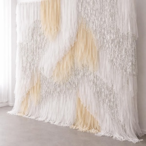 May include: A large decorative backdrop featuring layered vertical strips of white tulle, cream-coloured fabric, and shimmering silver metallic fringe. The materials are arranged in a diagonal, overlapping pattern that creates a textured, multi-layered appearance across the entire surface of the hanging display.
