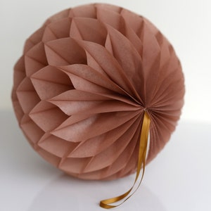 Pode incluir: Decoração esférica de papel em favo de mel em rosa suave. O papel é dobrado em um padrão repetitivo, criando uma superfície texturizada. Uma fita dourada é anexada para pendurar. Adequado para festas e decoração de casa.