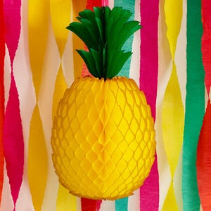 Può includere: Una grande decorazione di ananas di carta gialla con foglie di carta verde. L'ananas ha un design a nido d'ape. Lo sfondo presenta strisce verticali colorate in rosso, rosa, giallo e verde.