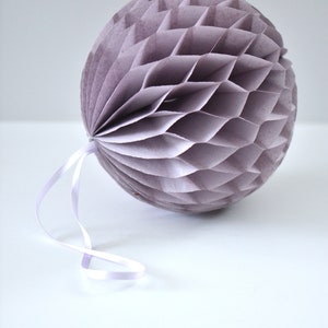 Pode incluir: Uma decoração esférica de papel em favo de mel num tom lavanda suave. A estrutura apresenta um padrão geométrico repetitivo criado por finas dobras de papel em camadas. Uma fita fina está presa ao topo para permitir a sua suspensão.