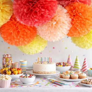 Peut inclure: Une scène de fête d'anniversaire avec un gâteau blanc orné de bougies, des cupcakes et des bonbons colorés. Au-dessus, des pompons en papier aux couleurs vives, orange, jaune et rose, créent une ambiance joyeuse. Des confettis parsèment la table.