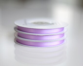 May include: A stack of three spools of light purple satin ribbon. Each spool features a smooth, shiny texture and is wound around a white circular cardboard core. The ribbon is uniform in color and width, creating a neat, layered appearance.