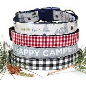 May include: A stack of four dog collars with various patterns. The top collar has a mountain and tree design with a blue buckle. The second collar has the text "ADVENTURE AWAITS". The third is red and white checkered. The bottom collar has the text "HAPPY CAMPER".