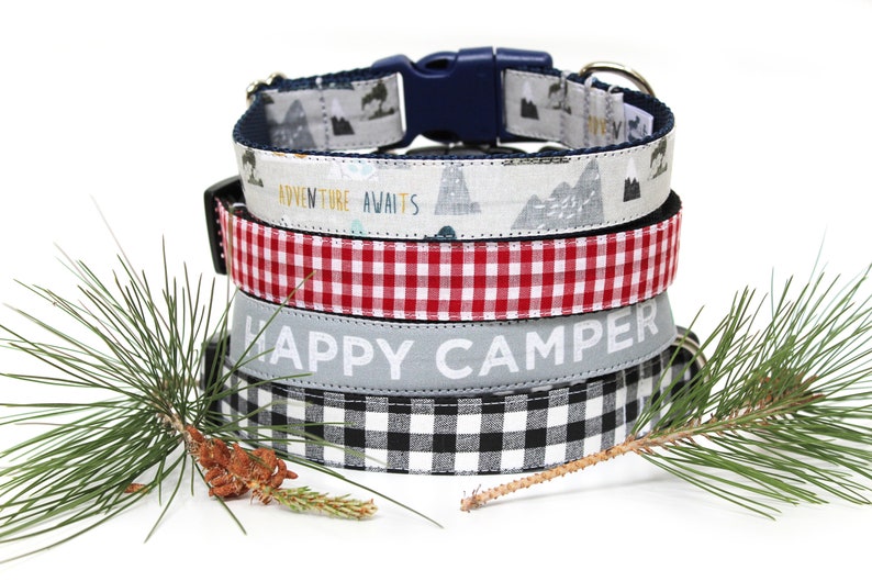 May include: A stack of four dog collars with various patterns. The top collar has a mountain and tree design with a blue buckle. The second collar has the text "ADVENTURE AWAITS". The third is red and white checkered. The bottom collar has the text "HAPPY CAMPER".