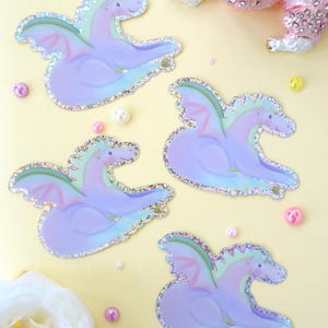 Peut inclure: Quatre autocollants de dragons violets pastel avec des ailes irisées et des bords scintillants sont disposés sur une surface jaune. Une figurine de caniche rose ornée de bijoux et des perles éparpillées complètent le thème fantaisiste. Une fleur blanche à cœur jaune est également présente.