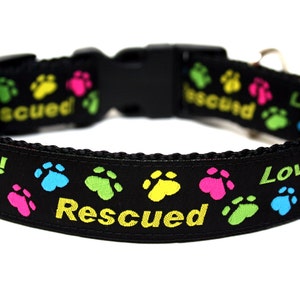 May include: Black dog collar with colorful paw prints and the words "Rescued" and "Loved!" in yellow and green. The collar has a black plastic buckle.